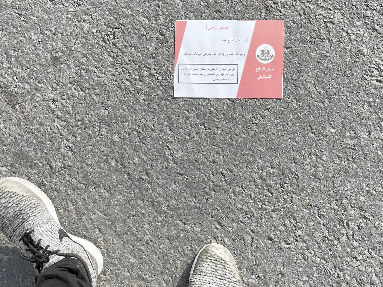 Ett rött och vitt flygblad med arabisk text och den israeliska arméns emblem ligger på asfalten framför två fötter i grå gymnastikskor.